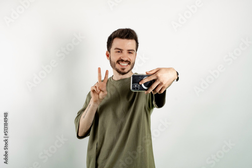 Cheerful caucasian man wearing casual clothes posing isolated over white background gesturing victory sign and smiling while taking selfie. Video call or social media concepts.