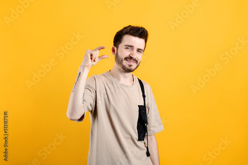 Caucasian man wearing beige tee posing isolated over yellow background looking at camera showing measurement with fingers. Showing small size length with a pity.