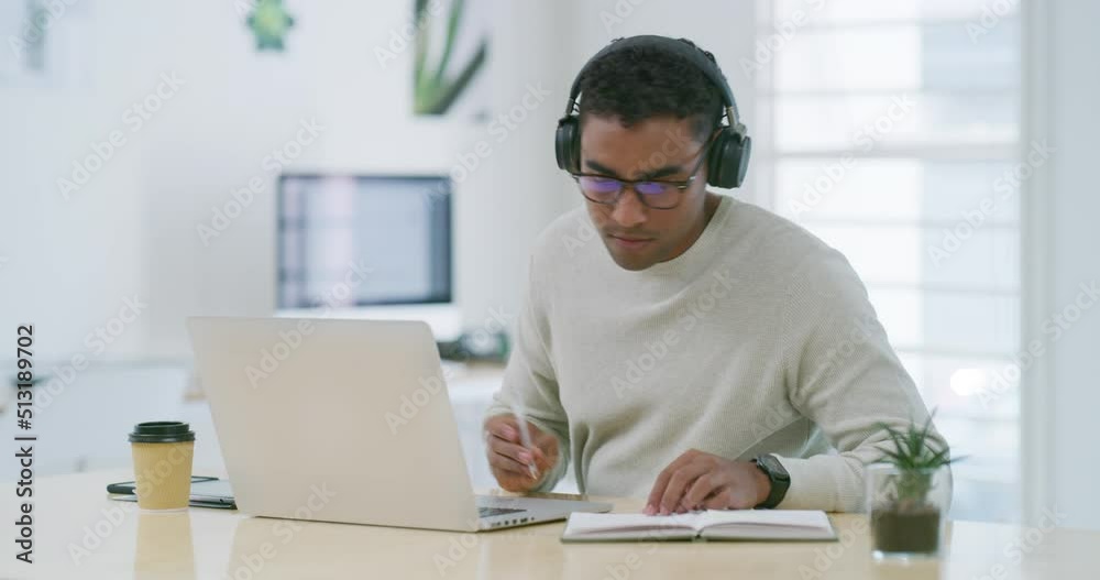 Mixed race creative entrepreneur using a laptop, writing notes in a notebook while listening to music on headphones. Young latino businessman and designer sitting alone and working from home office