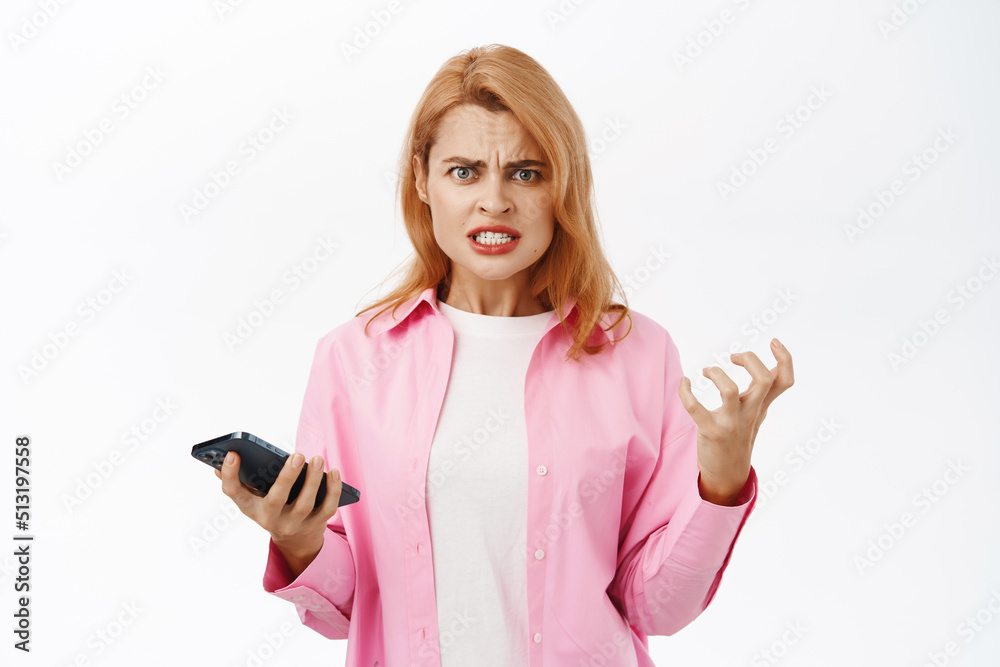 Girl holds mobile phone and boils from anger, shaking furious, stands over white background Girl holds mobile phone and boils from anger, shaking furious, stands over white background