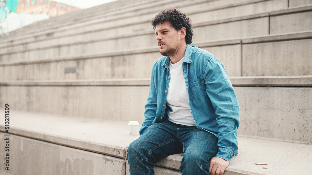 Upset man sits on the stairs and thinks emotionally. Closeup portrait of emotional young bearded man in denim shirt sits on high steps. Upset man sits on the stairs and thinks emotionally. Closeup portrait of emotional young bearded man in denim shirt sits on high steps.