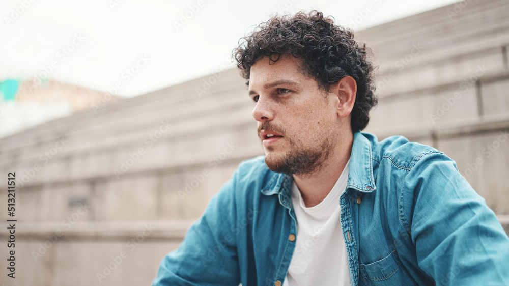 Upset man sits on the stairs and thinks emotionally. Closeup portrait of emotional young bearded man in denim shirt sits on high steps.