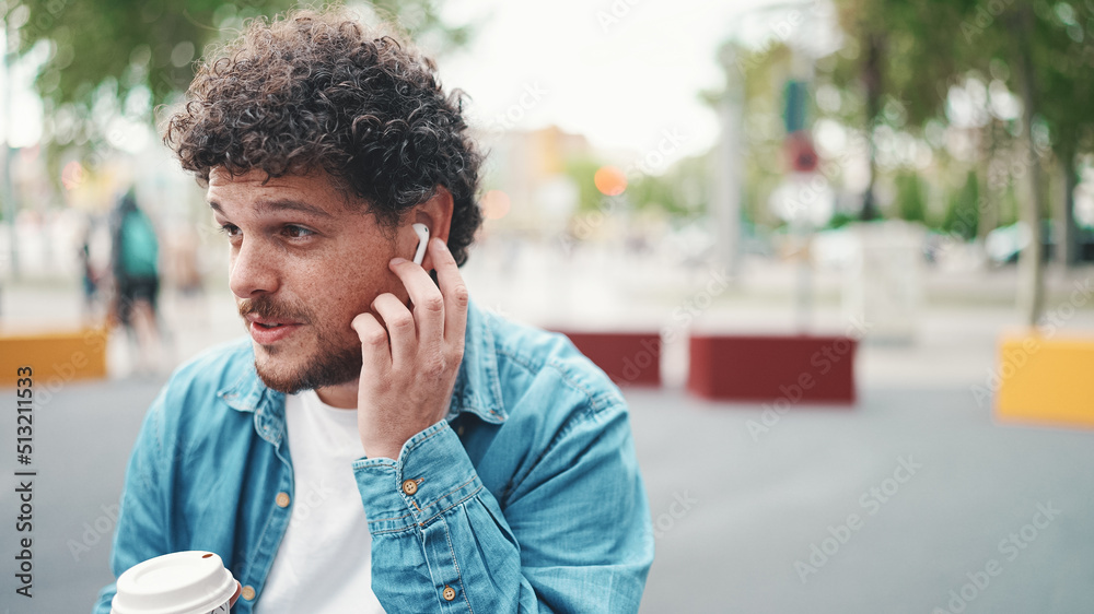 Close-up portrait of young bearded man in denim shirt sitting in wireless headphones and drinking coffee on busy street modern city background. Profile of man drinking coffee Close-up portrait of young bearded man in denim shirt sitting in wireless headphones and drinking coffee on busy street modern city background. Profile of man drinking coffee