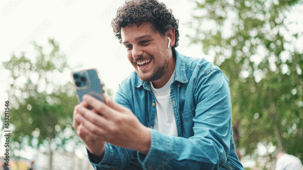 Clouseup, young bearded man in denim shirt sitting in wireless headphones making video call on mobile phone on busy street modern city background.