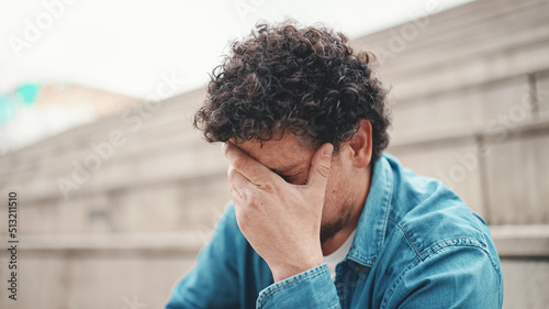 Upset man sits on the stairs and thinks emotionally. Closeup portrait of emotional young bearded man in denim shirt sits on high steps.