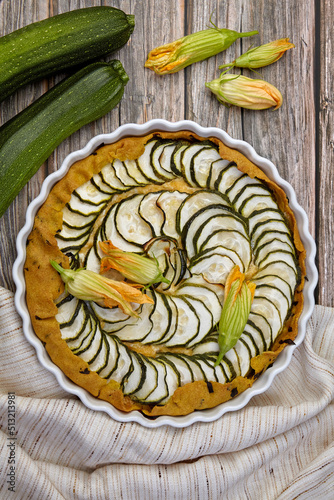 Delicious homemade zucchini pie with curd cheese and herbs surrounded by flowers, seeds and zucchin