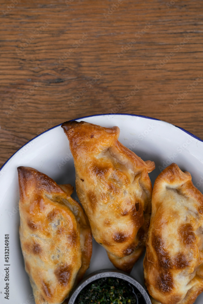 Traditional Argentinian food. Top view of empanadas with chimichurri dipping sauce, in a white bowl on the wooden table. Traditional Argentinian food. Top view of empanadas with chimichurri dipping sauce, in a white bowl on the wooden table.