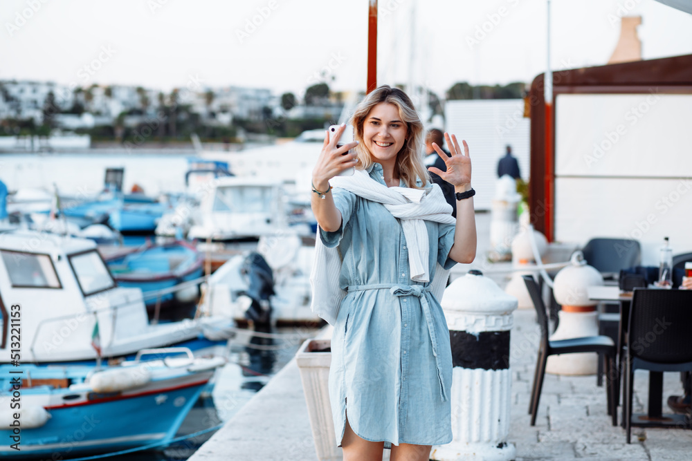 Beautiful young woman, denim dress takes selfie on shore. among yachts, on pier. full-length photo. happy holidays. Beautiful young woman, denim dress takes selfie on shore. among yachts, on pier. full-length photo. happy holidays.
