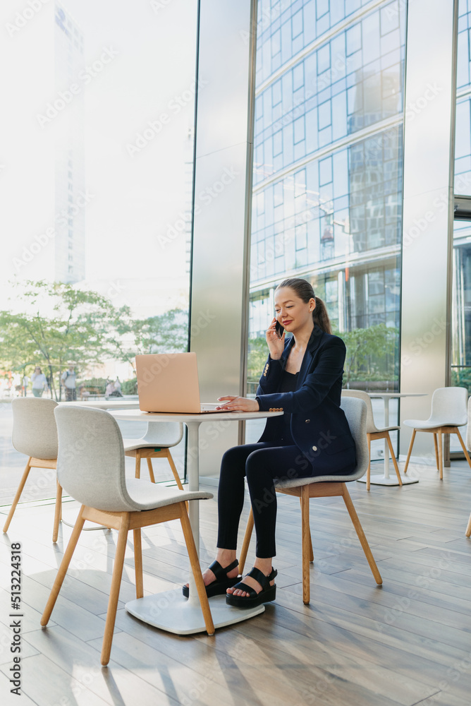A full length photo of a female realtor who is having conversation on phone. A happy woman is sitting at the table in the modern lobby. A female IT developer near a laptop in a contemporary interior.