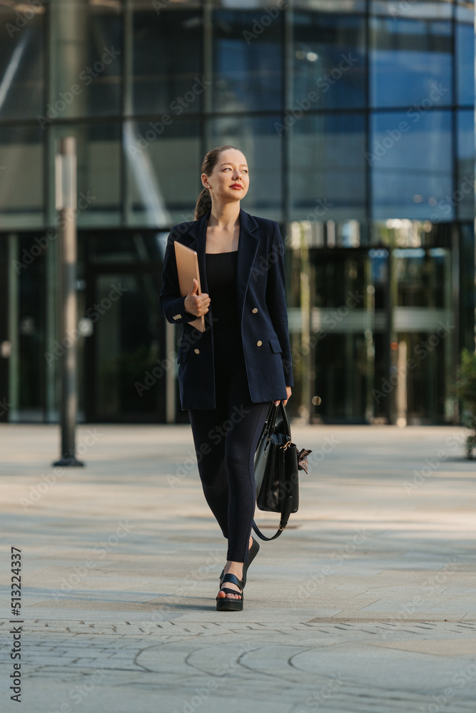 A full length photo of a female realtor in a blazer who is strolling with a laptop between skyscrapers.