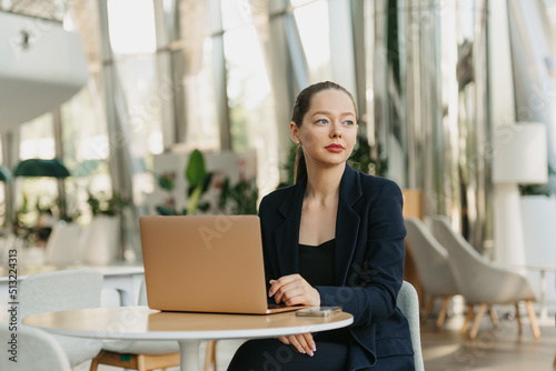 A female realtor in a blazer is waiting for a client. A woman is sitting at the table and working in the modern lobby. A female IT developer is relaxing near a laptop in a contemporary office interior