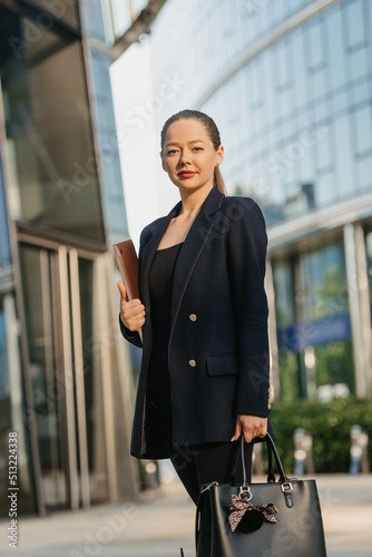 A female realtor in a blazer is posing with a laptop between skyscrapers. A businesswoman is leaving her office in a downtown in the evening.