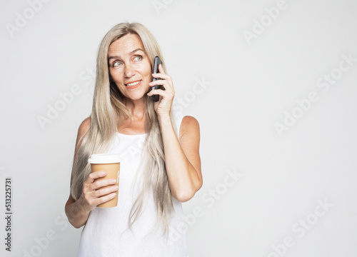 Happy elderly female with lomg hair makes mobile call, drinks takeaway coffee photo