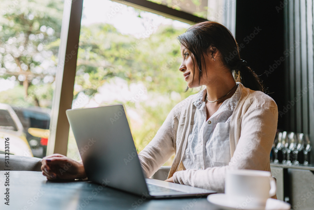 Nomad on work. Confident young Latin woman making video call on laptop while sitting by window in cafe. Nomad on work. Confident young Latin woman making video call on laptop while sitting by window in cafe.