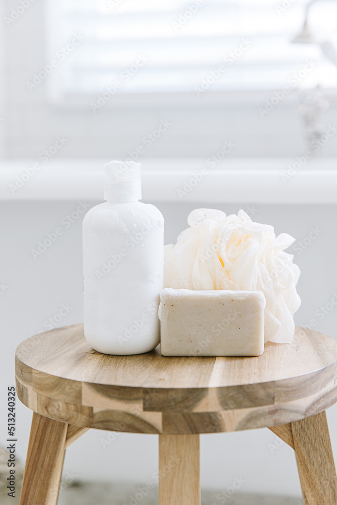 A white soap bottle with no label on it. There's also a bar of soap sitting there with a loofah sponge. All the products are on a wood stool. In the background is a bath tub and a window. A white soap bottle with no label on it. There's also a bar of soap sitting there with a loofah sponge. All the products are on a wood stool. In the background is a bath tub and a window.