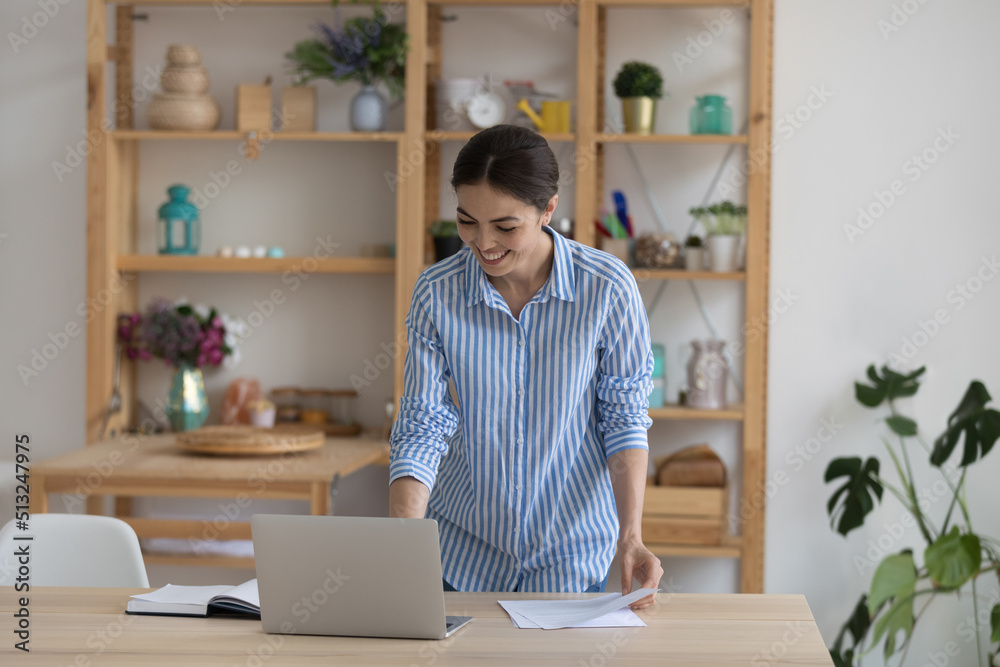 Smiling business lady standing at workplace finish urgent paperwork looks satisfied by result. Leader of project working in modern cozy office use laptop, sorts out papers feel happy. Workflow concept Smiling business lady standing at workplace finish urgent paperwork looks satisfied by result. Leader of project working in modern cozy office use laptop, sorts out papers feel happy. Workflow concept