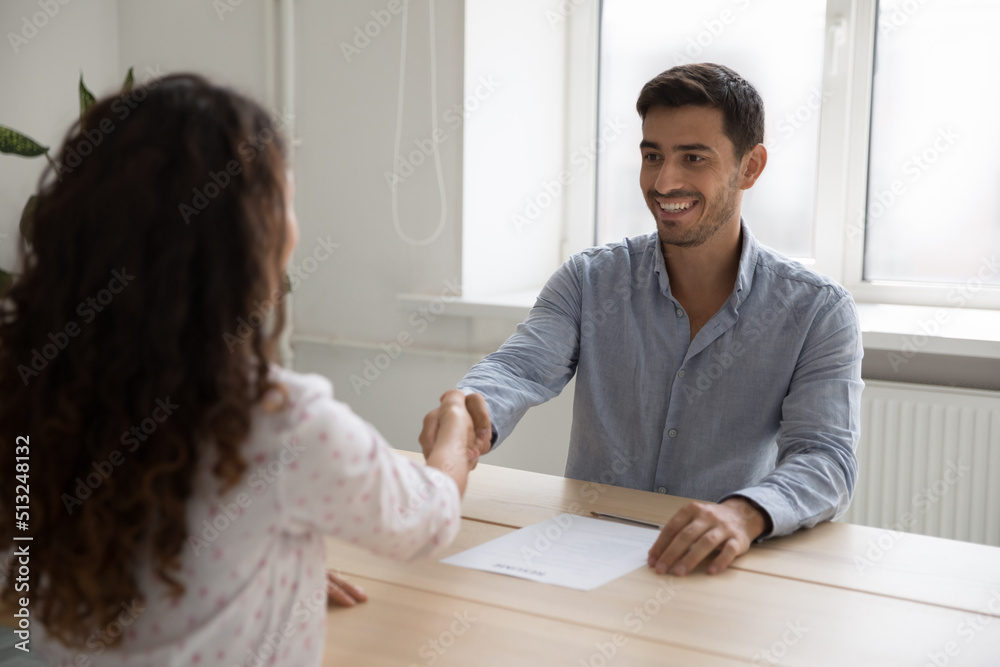 Human resources, hiring, headhunt concept. Hispanic businessman shake hands female applicant sit at desk during successful job interview, making commercial deal, sign contract, buy services of company