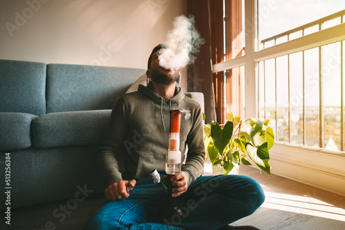 Man smoking bong and exhaling the smoke at home. Man smoking pot, medical marijuana or cannabis from a bong or water pipe. Cannabis and weed legalisation concept