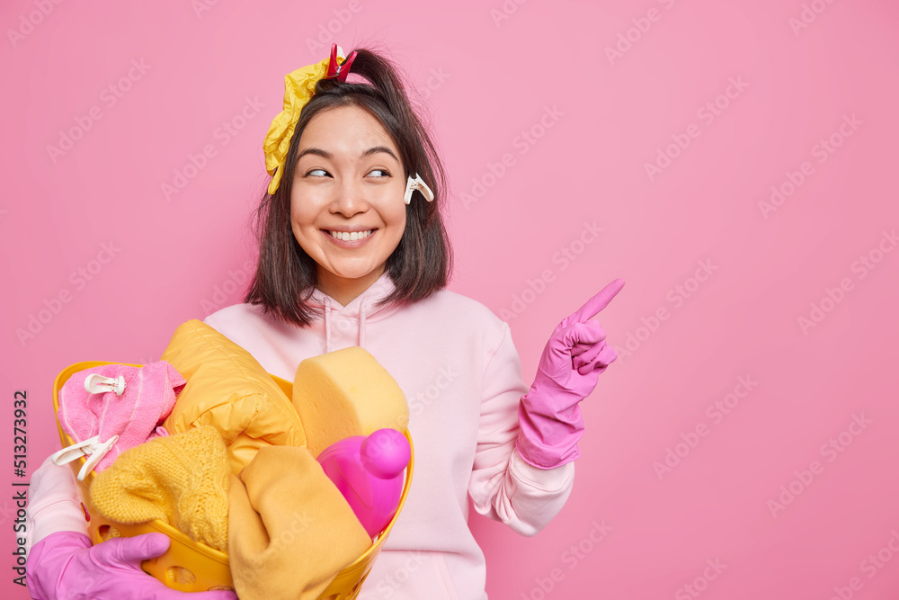Cheerful housewife smiles gladfully points right on blank space advertises something carries basin with laundry and detergents isolated over pink background. Routine household duties concept Cheerful housewife smiles gladfully points right on blank space advertises something carries basin with laundry and detergents isolated over pink background. Routine household duties concept