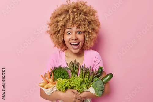 Happy surprised woman likes grocery shopping holds variety of healthy vegetables looks with wonder keeps to nutritionn for weight loss stands against pink background. Vegeterian food concept photo