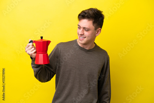 Young Brazilian man holding coffee pot isolated on yellow background looking to the side and smiling