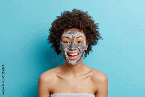 Indoor shot of glad young woman with curly dark hair smiles happily applies beauty clay mask on face undergoes skin care cosmetic procedures stands bare shoulders against blue studio background. photo