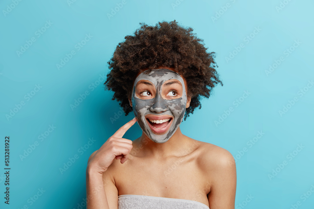 Cheerful woman with curly hair dreamy expression points at face undergoes anit aging skin care procedures applies clay mask stands wrapped in towel isolated over blue background. Beauty concept Cheerful woman with curly hair dreamy expression points at face undergoes anit aging skin care procedures applies clay mask stands wrapped in towel isolated over blue background. Beauty concept
