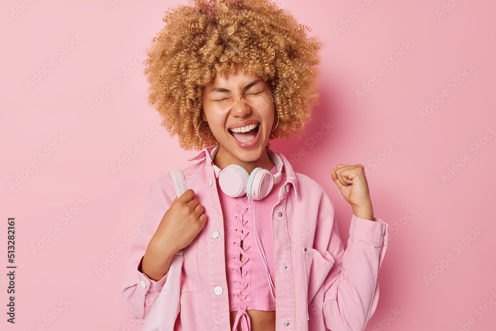 Positive woman with curly hair clenches fist celebrates success feels like winner dressed in jacket carries fabric bag uses stereo headphones isolated over pink background. Student glad to pass exams Positive woman with curly hair clenches fist celebrates success feels like winner dressed in jacket carries fabric bag uses stereo headphones isolated over pink background. Student glad to pass exams