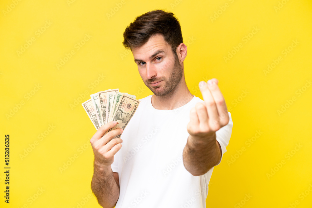 Young caucasian man taking a lot of money isolated on yellow background making money gesture Young caucasian man taking a lot of money isolated on yellow background making money gesture