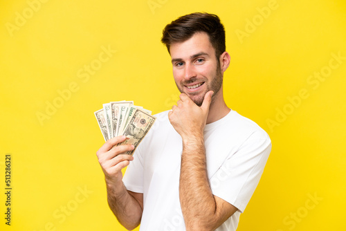 Young caucasian man taking a lot of money isolated on yellow background happy and smiling