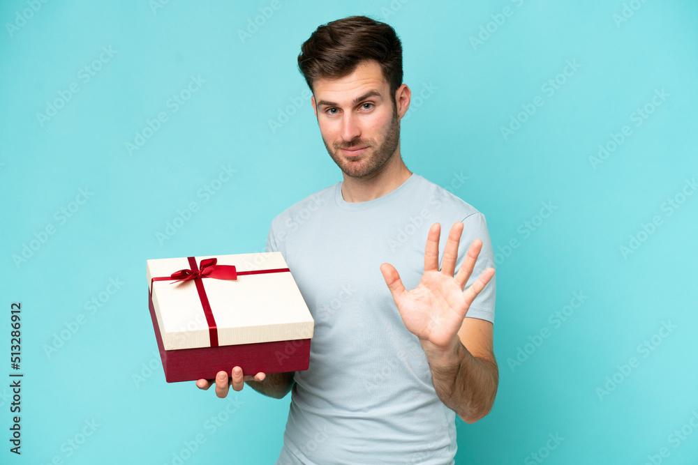 Young caucasian man holding a gift isolated on blue background saluting with hand with happy expression Young caucasian man holding a gift isolated on blue background saluting with hand with happy expression