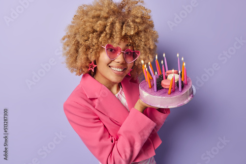 Happy fashionable woman wears trendy sunglasses and pink formal jacket holds sweet cake with burning candles going to make wish on her birthday isolated over purple background. Celebration concept photo