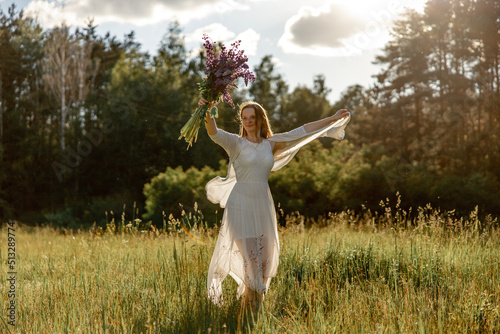 Young beautiful woman, wearing white dress, holding flowers and dancing on the meadow. Girl joying nature and freedom. Natural beauty. Dance, movement. Mental health, stress free, dreaming. Sunset.