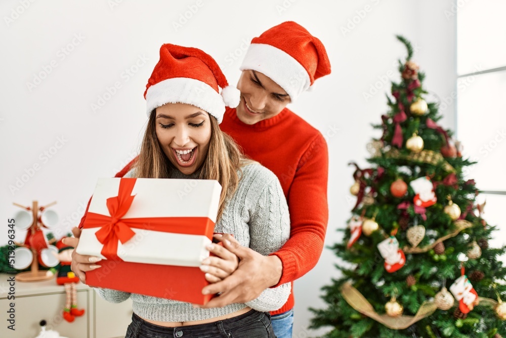 Young couple hugging and smiling happy. Wearing christmas hat holding gift at home. Young couple hugging and smiling happy. Wearing christmas hat holding gift at home.