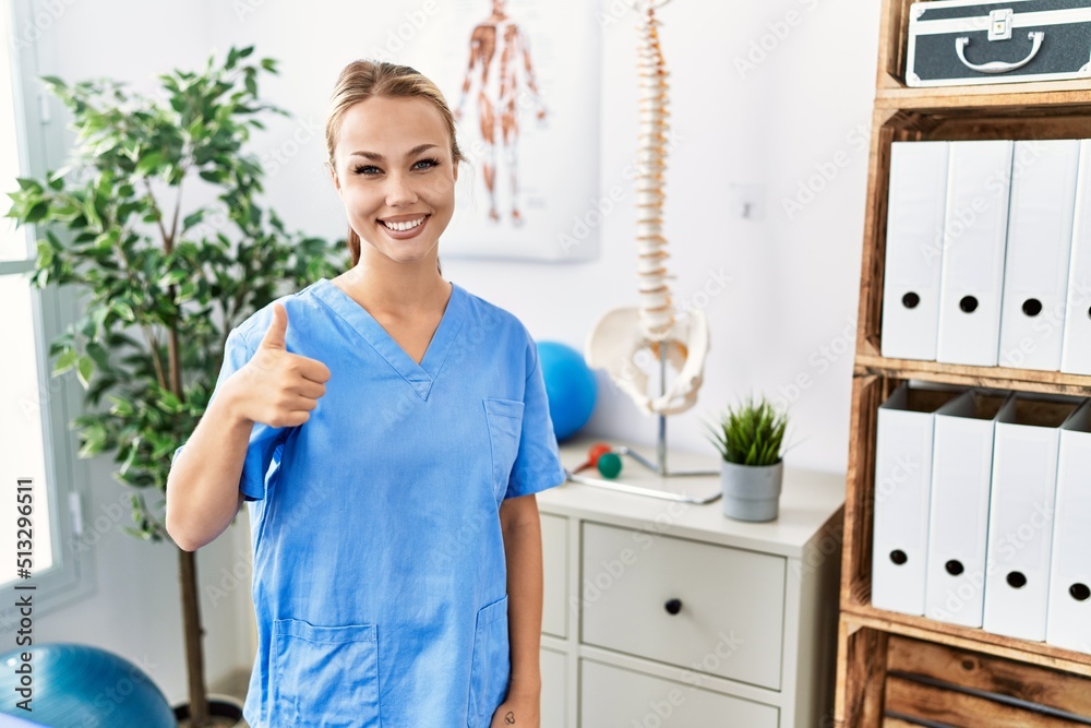 Young caucasian woman working at pain recovery clinic doing happy thumbs up gesture with hand. approving expression looking at the camera showing success. Young caucasian woman working at pain recovery clinic doing happy thumbs up gesture with hand. approving expression looking at the camera showing success.
