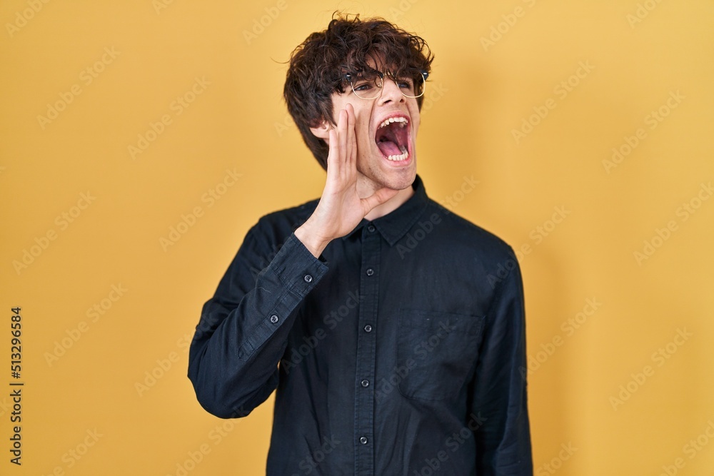 Young man wearing glasses over yellow background shouting and screaming loud to side with hand on mouth. communication concept.