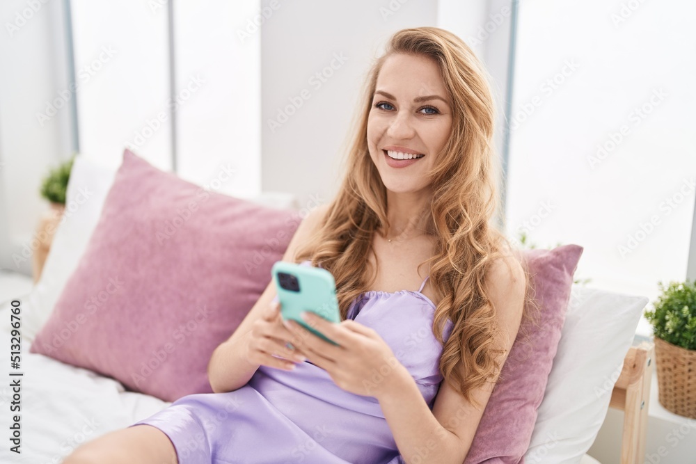Young blonde woman using smartphone sitting on bed at bedroom Young blonde woman using smartphone sitting on bed at bedroom
