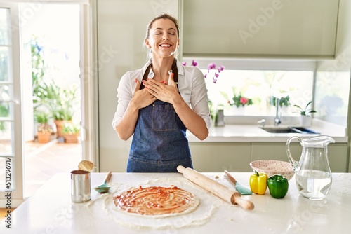 Beautiful blonde woman wearing apron cooking pizza smiling with hands on chest with closed eyes and grateful gesture on face. health concept.