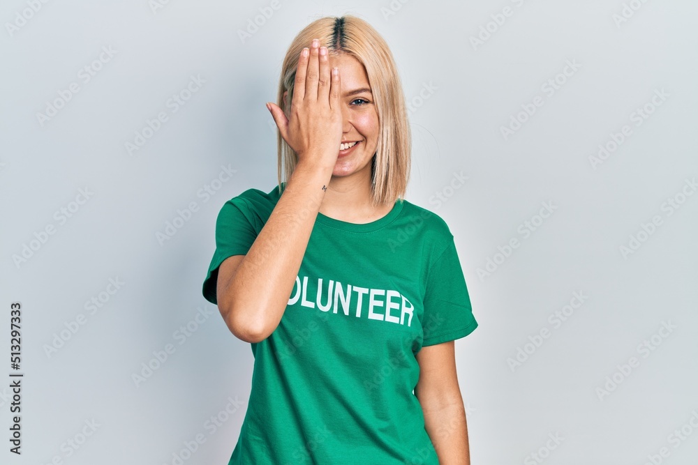 Beautiful blonde woman wearing volunteer t shirt covering one eye with hand, confident smile on face and surprise emotion. Beautiful blonde woman wearing volunteer t shirt covering one eye with hand, confident smile on face and surprise emotion.