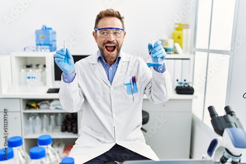 Middle age man working at scientist laboratory holding chemical products smiling and laughing hard out loud because funny crazy joke.