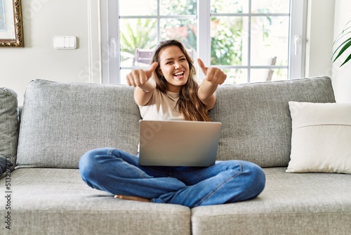 Fotografi Beautiful young brunette woman sitting on the sofa using computer laptop at home approving doing positive gesture with hand, thumbs up smiling and happy for success