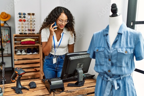 Young latin shopkeeper woman talking on the smartphone working at clothing store.