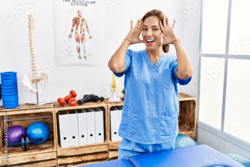 Middle age hispanic physiotherapist woman working at pain recovery clinic smiling cheerful playing peek a boo with hands showing face. surprised and exited