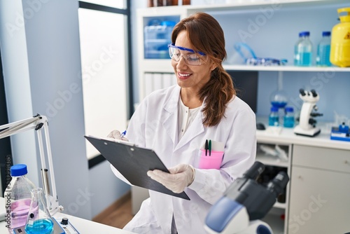 Young latin woman wearing scientist uniform writing on clipboard at laboratory