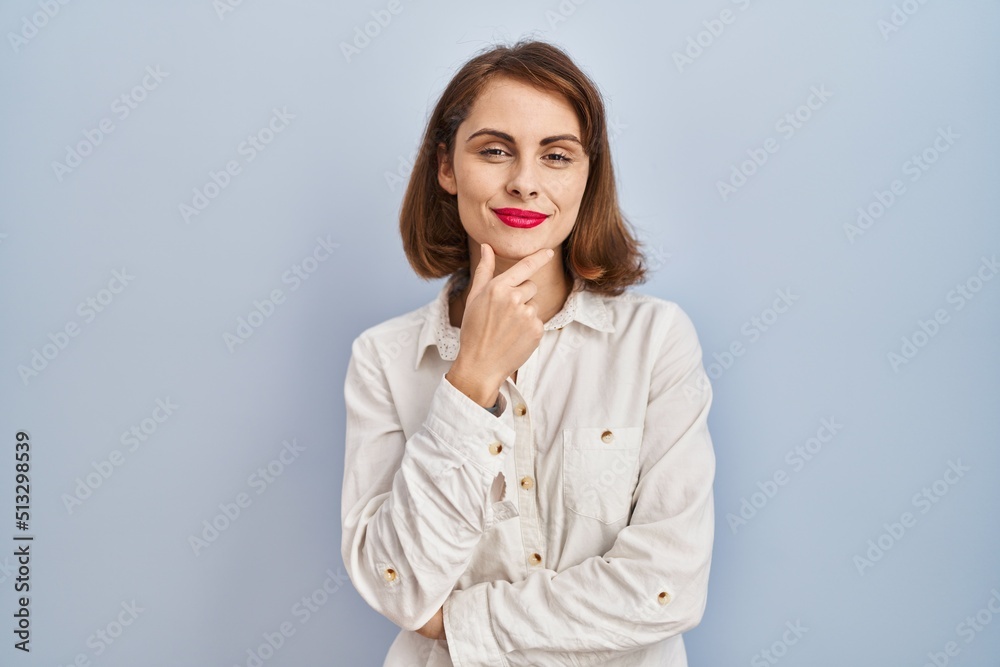 Young beautiful woman standing casual over blue background looking confident at the camera with smile with crossed arms and hand raised on chin. thinking positive. Young beautiful woman standing casual over blue background looking confident at the camera with smile with crossed arms and hand raised on chin. thinking positive.