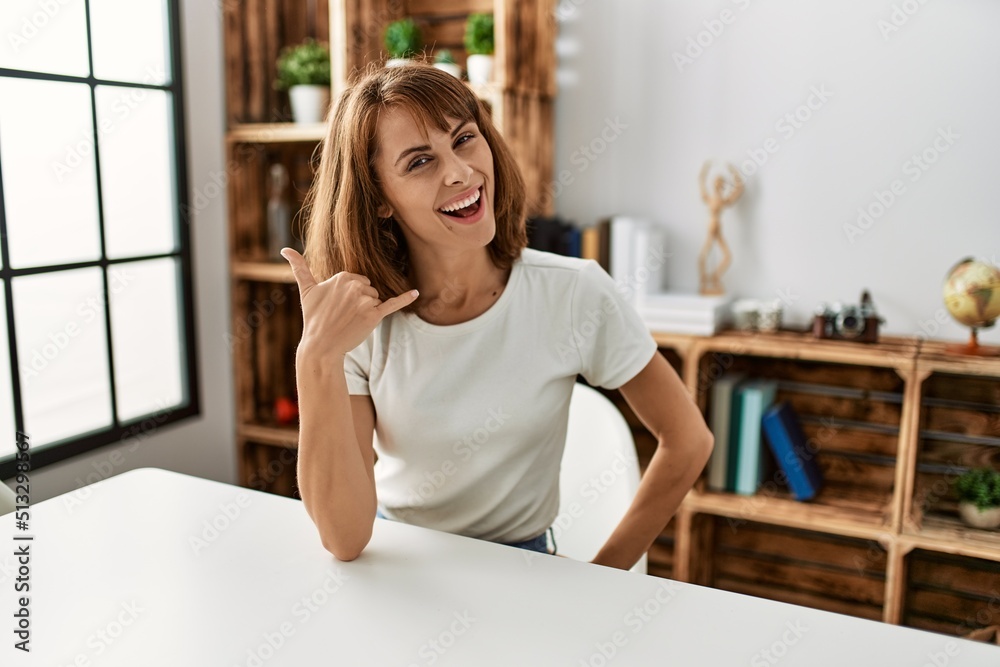 Young caucasian girl wearing casual clothes sitting on the table at home smiling doing phone gesture with hand and fingers like talking on the telephone. communicating concepts. Young caucasian girl wearing casual clothes sitting on the table at home smiling doing phone gesture with hand and fingers like talking on the telephone. communicating concepts.