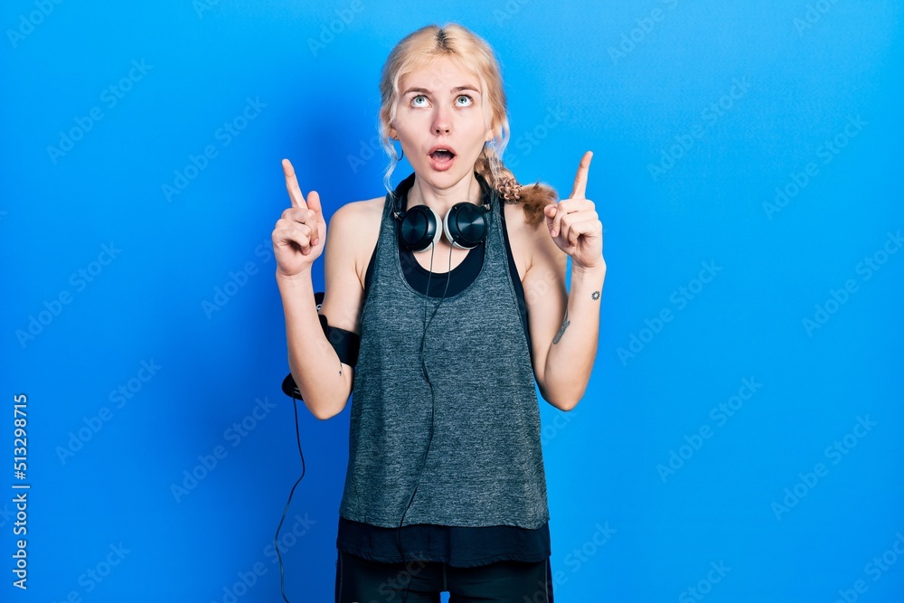 Fototapeta premium Beautiful caucasian woman with blond hair wearing sportswear amazed and surprised looking up and pointing with fingers and raised arms.