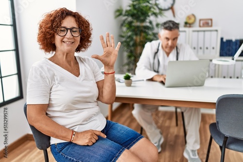 Senior woman sitting at doctor appointment showing and pointing up with fingers number five while smiling confident and happy.