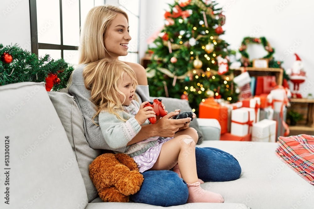 Mother and daughter playing video game sitting by christmas tree at home