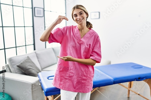 Young blonde woman working as physiotherapist at home gesturing with hands showing big and large size sign  measure symbol. smiling looking at the camera. measuring concept.